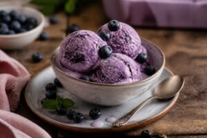 Cremiges Heidelbeer-Vanilleeis ohne Eismaschine in einer Glasschale, garniert mit frischen Blaubeeren und Vanillemark, auf einem hellen Holztisch