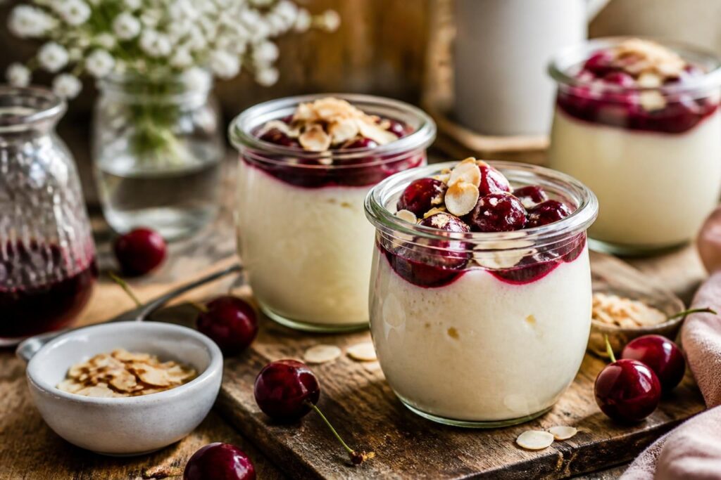 Kirsch-Mandel-Pudding im Glas mit Sauerkirschen, Mandelcreme und gehobelten Mandeln – cremiges Kühlschrank-Dessert in vier Portionsgläsern auf einem Holztablett