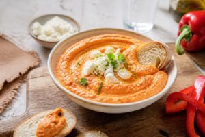 Kalte Paprika-Feta-Creme in einer weißen Schüssel, garniert mit Olivenöl und frischen Kräutern, serviert mit Fladenbrot und Gemüsesticks