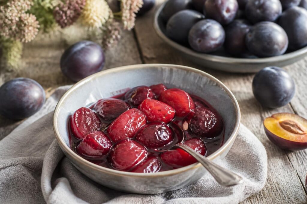 Pflaumenröster ohne Gelierzucker in einem hellen Keramiktopf, ganze Zwetschgenstücke in dunkelrotem Sirup, mit Zimtstange und Sternanis garniert