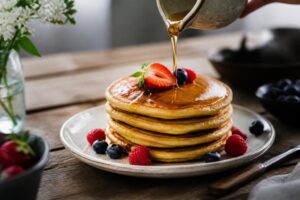 Fluffige amerikanische Pancakes gestapelt auf einem weißen Teller, mit Ahornsirup und frischen Beeren garniert, Draufsicht auf Holzuntergrund